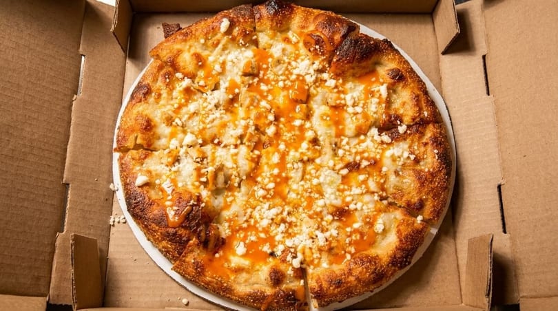 Buffalo chicken pizza with crumbled cheese and orange sauce in a cardboard delivery box.