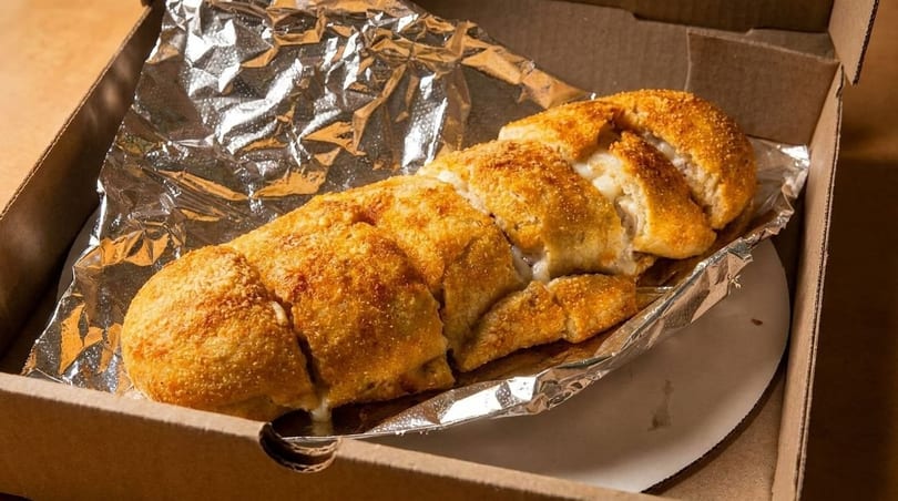 Golden-brown cheesy breadsticks, sliced and oozing with white cheese, in a cardboard box lined with foil.